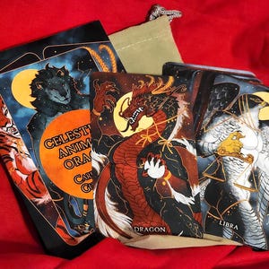 May include: A set of Celestial Animal Oracle Cards with a black background and gold accents. The cards feature illustrations of animals in a celestial setting. The cards are arranged in a fan shape, with the top card showing a dragon.