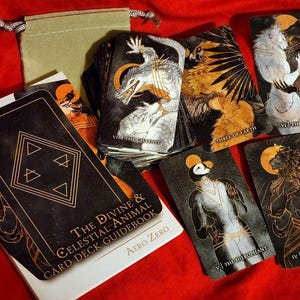 Puede incluir: Una baraja de tarot negra y dorada con un libro de gu&iacute;a. Las cartas presentan ilustraciones de animales e im&aacute;genes celestiales. El libro de gu&iacute;a tiene el t&iacute;tulo "The Divine & Celestial Animal Card Deck Guidebook" y el autor "Aero Zero".