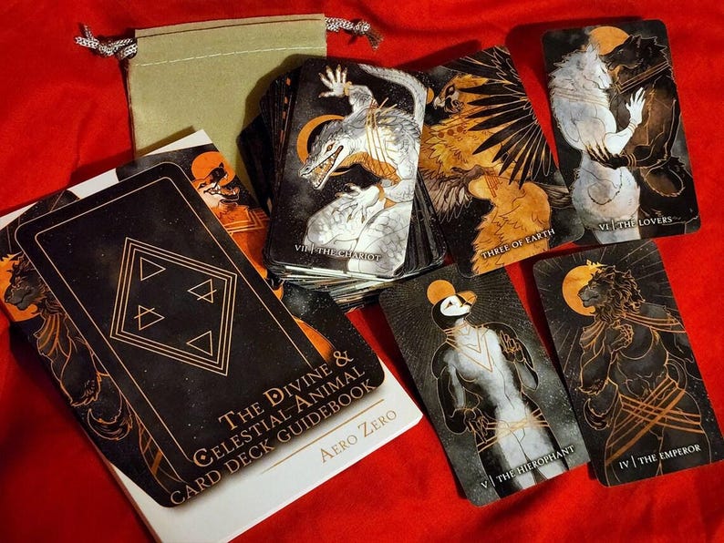 Puede incluir: Una baraja de tarot negra y dorada con un libro de gu&iacute;a. Las cartas presentan ilustraciones de animales e im&aacute;genes celestiales. El libro de gu&iacute;a tiene el t&iacute;tulo "The Divine & Celestial Animal Card Deck Guidebook" y el autor "Aero Zero".