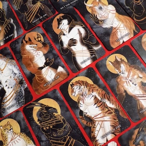 Puede incluir: Un juego de cartas de tarot con ilustraciones de animales antropomorfos en varias poses. Las cartas est&aacute;n decoradas con detalles dorados y tienen un fondo negro con un patr&oacute;n estrellado. Las cartas incluyen los siguientes t&iacute;tulos: "Four of Fire", "Eight of Air", "XII The Hang", "Queen of Air", "Seven of Air", "Page of Air", "Knight of Fire" y "Three of Fire".
