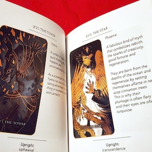 Puede incluir: Dos cartas de tarot, XVI: La Torre y XVII: La Estrella, con ilustraciones de una hiena y un f&eacute;nix. La carta de la Torre presenta una hiena negra y dorada con una expresi&oacute;n feroz. La carta de la Estrella presenta un f&eacute;nix blanco con plumas doradas y naranjas, sobre un fondo negro.