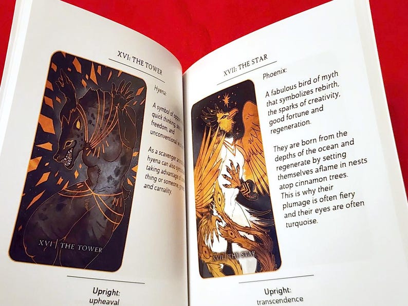 Puede incluir: Dos cartas de tarot, XVI: La Torre y XVII: La Estrella, con ilustraciones de una hiena y un f&eacute;nix. La carta de la Torre presenta una hiena negra y dorada con una expresi&oacute;n feroz. La carta de la Estrella presenta un f&eacute;nix blanco con plumas doradas y naranjas, sobre un fondo negro.