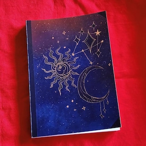 Pu&ograve; includere: Un taccuino con un design celeste con sole, luna e stelle su sfondo blu scuro. La copertina presenta illustrazioni color oro e un dorso nero. Il taccuino &egrave; posto su uno sfondo rosso.