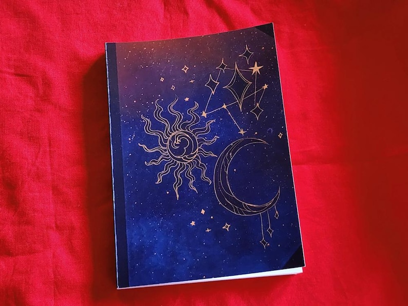 Pu&ograve; includere: Un taccuino con un design celeste con sole, luna e stelle su sfondo blu scuro. La copertina presenta illustrazioni color oro e un dorso nero. Il taccuino &egrave; posto su uno sfondo rosso.