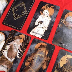 Puede incluir: Un juego de cartas de tarot con ilustraciones de animales antropomorfos. Las cartas son negras con detalles dorados y presentan una variedad de animales, incluyendo un lobo, un p&aacute;jaro, un ciervo y un oso. Las cartas est&aacute;n dispuestas en forma de abanico sobre una superficie roja. Las cartas tienen t&iacute;tulos como "Rey del Agua", "Diez del Aire" y "Caballero del Aire".
