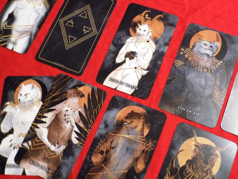 Puede incluir: Un juego de cartas de tarot con ilustraciones de animales antropomorfos. Las cartas son negras con detalles dorados y presentan una variedad de animales, incluyendo un lobo, un p&aacute;jaro, un ciervo y un oso. Las cartas est&aacute;n dispuestas en forma de abanico sobre una superficie roja. Las cartas tienen t&iacute;tulos como "Rey del Agua", "Diez del Aire" y "Caballero del Aire".