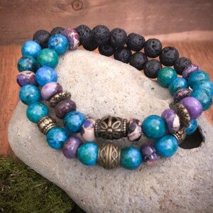 May include: A double-strand bracelet with blue, green, and purple beads, accented with gold-toned metal spacers and a larger, ornate metal charm. The bracelet is made with lava rock and gemstone beads.