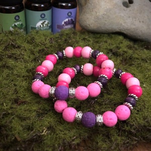 Pode incluir: Duas pulseiras de contas cor-de-rosa e roxas com detalhes prateados. As pulseiras são feitas de contas de rocha vulcânica e são projetadas para conter óleos essenciais.
