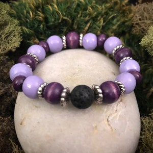 May include: A purple and lavender wooden bead bracelet with a black lava rock bead. The bracelet has silver accents on each bead.