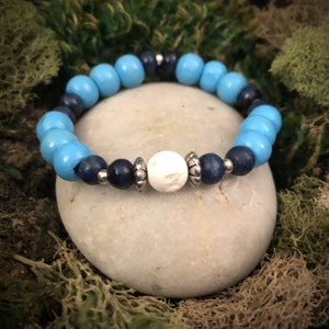 Könnte beinhalten: Ein blaues und dunkelblaues Perlenarmband mit einer weißen Perle in der Mitte. Das Armband liegt auf einem grauen Stein.