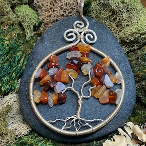 May include: A silver wire wrapped tree of life pendant with carnelian, agate, and clear quartz stones. The pendant is on a gray stone background.