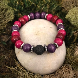 Puede incluir: Un brazalete hecho de cuentas de madera en tonos de rosa y morado con detalles plateados. El brazalete tiene una cuenta de lava negra en el centro.