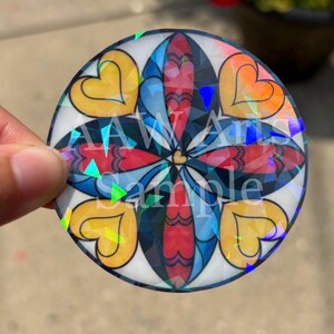 May include: A circular sticker featuring a symmetrical mandala-style design with four yellow hearts, red and blue petal shapes, and a central gold heart. The surface has a holographic finish with the text AAW Arts Sample overlaid in white.