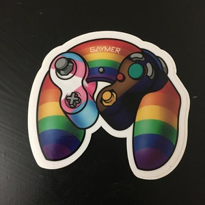 May include: A rainbow-colored GameCube controller with a transgender pride flag on the left controller and the word "GAYMER" in white text on the top of the controller.
