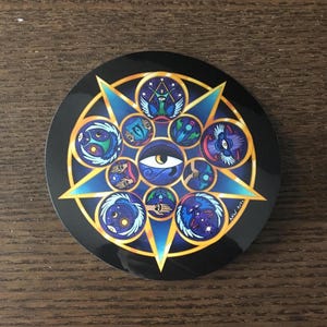 May include: Round sticker with a black background and a central design featuring a large eye surrounded by a starburst pattern. The design incorporates blue, gold, and teal elements, with various symbolic illustrations.
