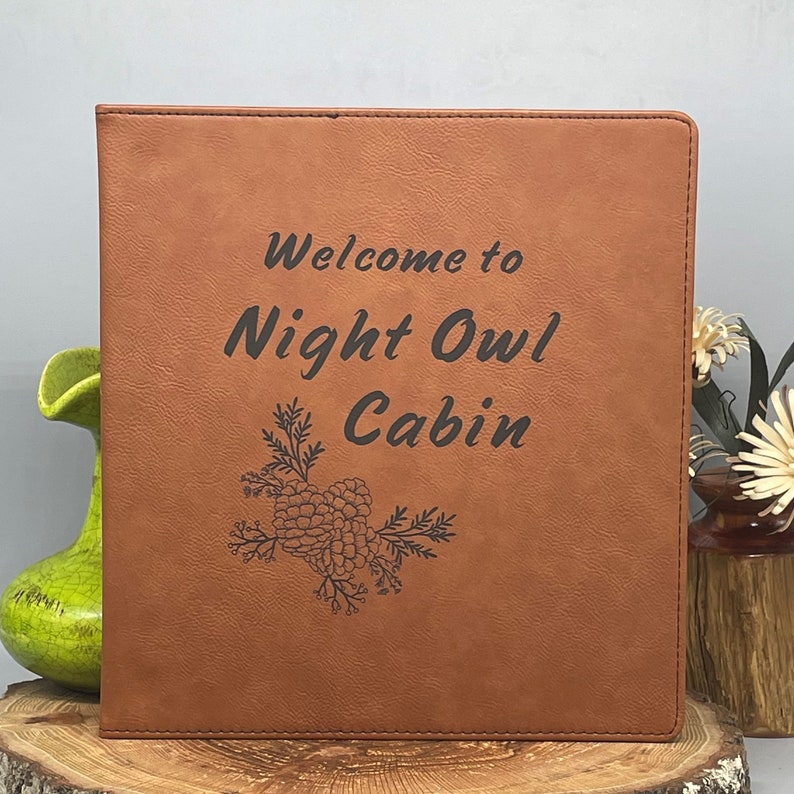 Peut inclure: Un journal reli&eacute; en cuir marron avec le texte "Welcome to Night Owl Cabin" et un motif de pomme de pin.