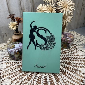 May include: A teal blue journal with a black silhouette of a dancer and the letter "S" surrounded by flowers. The name "Sarah" is printed in black at the bottom.