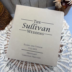 May include: A light beige wedding album with the text "The Sullivan Wedding" in black. The album includes the names Elaina and Connor, the date 9/12/2025, and the officiant's name, Christian Mikulski.