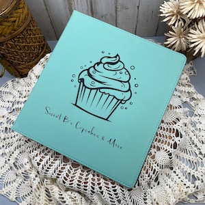 May include: A square, light blue binder featuring a black graphic of a cupcake with frosting and sprinkles. The text "Sweet B's Cupcakes & More" is printed in black below the cupcake illustration. The binder has stitched edges and a smooth finish.