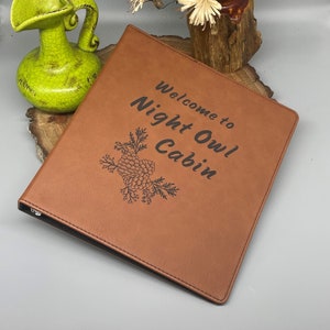 Peut inclure: Un classeur en cuir marron avec le texte "Bienvenue au Night Owl Cabin" et un motif de pomme de pin grav&eacute; sur le devant.