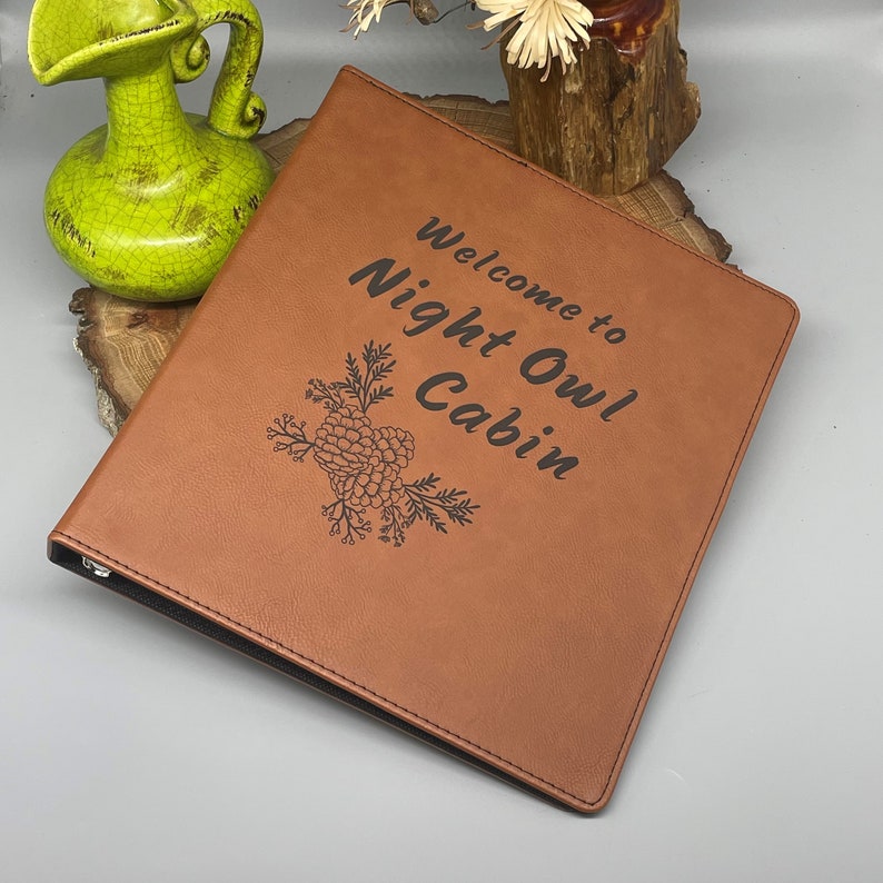Peut inclure: Un classeur en cuir marron avec le texte "Bienvenue au Night Owl Cabin" et un motif de pomme de pin grav&eacute; sur le devant.