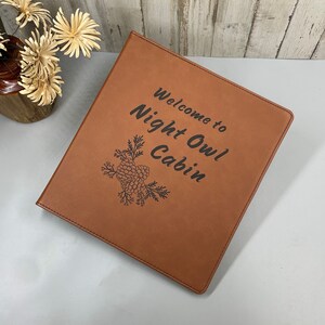 Peut inclure: Un classeur en cuir marron avec le texte "Welcome to Night Owl Cabin" et un motif de pomme de pin grav&eacute; sur le devant.