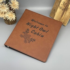 Peut inclure: Un classeur en cuir marron avec le texte "Welcome to Night Owl Cabin" et un motif de pomme de pin grav&eacute; sur le devant.