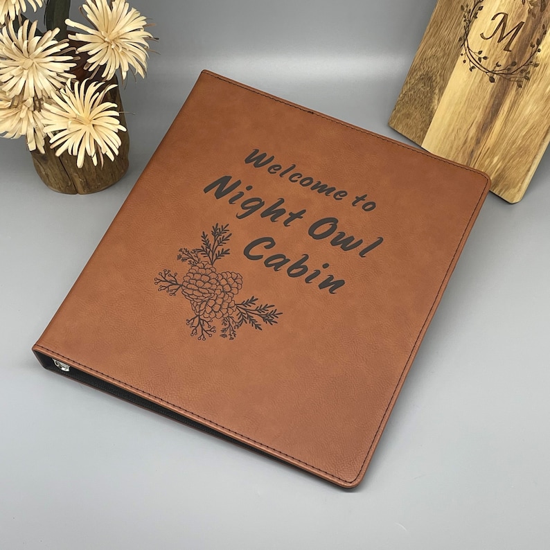 Peut inclure: Un classeur en cuir marron avec le texte "Welcome to Night Owl Cabin" et un motif de pomme de pin grav&eacute; sur le devant.