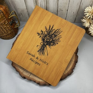 May include: A rectangular light brown bamboo recipe binder featuring a black laser-engraved illustration of a wildflower bouquet and the text "Sarah & Nick's Recipes" centered on the front cover with stitched edges along the perimeter.