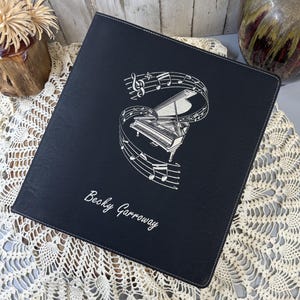 May include: Black leather binder with a silver embossed design of a grand piano and musical notes. The binder is personalized with the name "Becky Garroway".