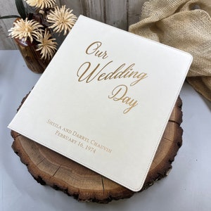 May include: A square white photo album with gold foil lettering on the cover that reads Our Wedding Day, followed by the names Sheila and Darryl Chauvin and the date February 16, 1974. The album has a smooth, leather-like texture and stitched edges.