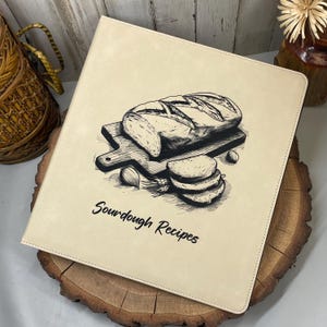May include: A beige recipe binder with a black and white illustration of a loaf of bread, slices, and garlic on a cutting board. The words "Sourdough Recipes" are printed in black script below the illustration. The binder is on a wooden surface.