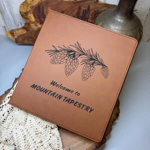 Peut inclure: Un journal reli&eacute; en cuir marron avec le texte "Bienvenue &agrave; Mountain Tapestry" et une silhouette noire de branches et de c&ocirc;nes de pin.