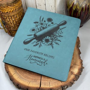 May include: A teal square recipe binder featuring a dark engraved illustration of a rolling pin surrounded by wildflowers. The text reads "OUR FAVORITE RECIPES" and "HAPPINESS IS Homemade" in a mix of block and script lettering on the front cover.