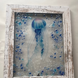 May include: A framed glass art piece featuring a blue jellyfish with a white background and blue glass pieces scattered around it.