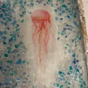May include: A pink jellyfish with long, flowing tentacles is depicted against a background of blue and green glitter. The jellyfish is the focal point of the image, with the glitter creating a shimmering effect.