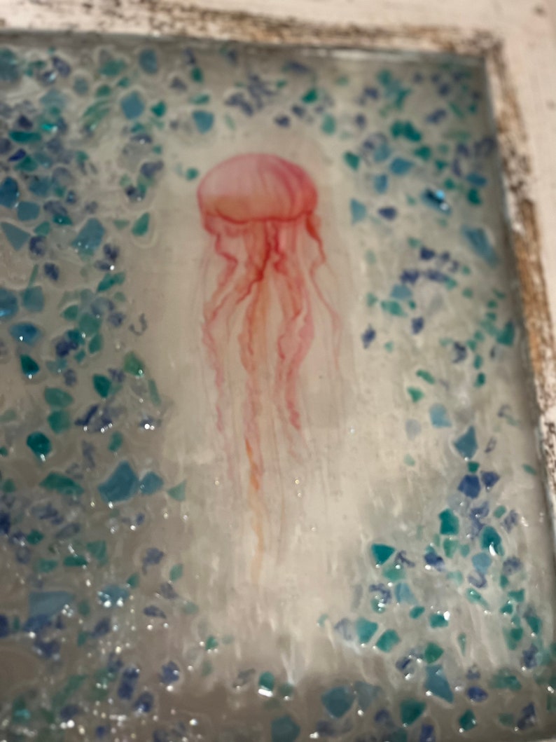 May include: A pink jellyfish with long, flowing tentacles is depicted against a background of blue and green glitter. The jellyfish is the focal point of the image, with the glitter creating a shimmering effect.