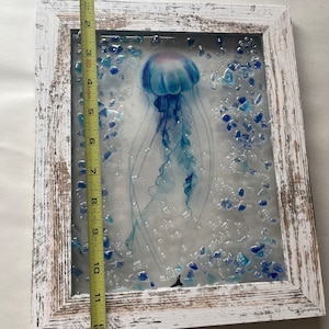 May include: A white wooden frame with a glass panel featuring a blue jellyfish with a light blue and purple gradient. The jellyfish is surrounded by small blue glass pieces and white dots.