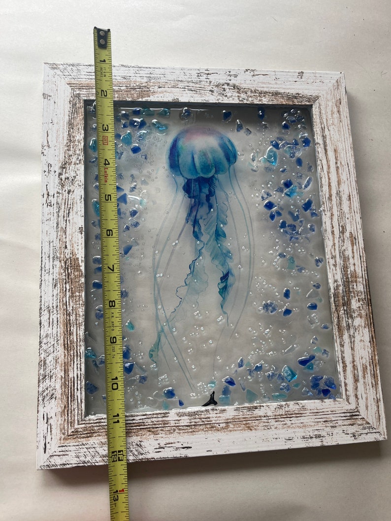 May include: A white wooden frame with a glass panel featuring a blue jellyfish with a light blue and purple gradient. The jellyfish is surrounded by small blue glass pieces and white dots.