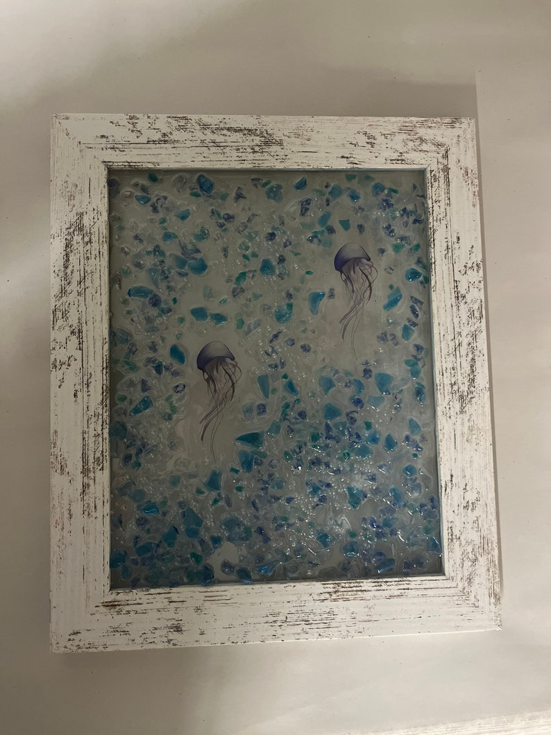 May include: A white framed picture with a blue and green background of small glass pieces. Two purple jellyfish are depicted in the picture.
