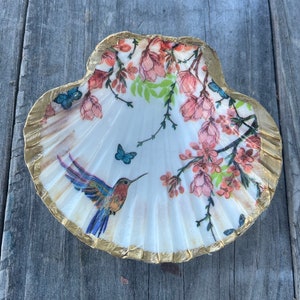 May include: A decorative seashell dish with a gold-painted rim. The interior features a hummingbird, butterflies, and floral design in shades of pink, blue, and green. The dish is resting on a weathered wooden surface.