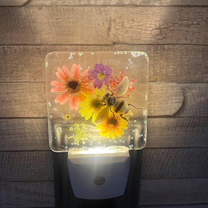 May include: A decorative night light featuring a square resin panel with embedded flowers and a bee. The flowers are pink, yellow, and purple. The night light has a white base and emits a soft glow.