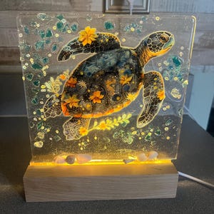 May include: A decorative illuminated resin lamp featuring a sea turtle design. The turtle is adorned with orange and yellow floral accents. The lamp sits on a wooden base and has a warm, inviting glow. The lamp is approximately 8 inches tall.