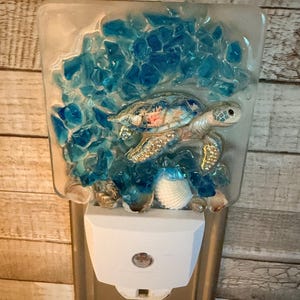 May include: A square glass night light featuring a sea turtle figurine surrounded by blue glass shards and small seashells. The turtle has a patterned shell with pink and white accents. The night light is mounted on a white plastic plug-in base.
