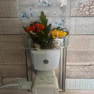 May include: A decorative night light featuring a clear, rectangular resin design with two small, colorful fish, green seaweed, and blue glass beads. The night light plugs into a wall outlet and has a white base.
