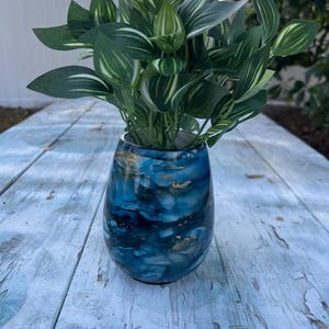 May include: A decorative vase with a blue and gold abstract design holds a bundle of green and white striped artificial leaves. The vase is on a weathered, light blue wooden surface.