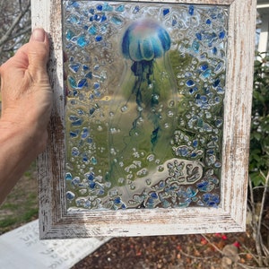 May include: A framed glass art piece featuring a blue jellyfish with a pink bell, surrounded by blue and clear glass pieces. The frame is white with a distressed finish.