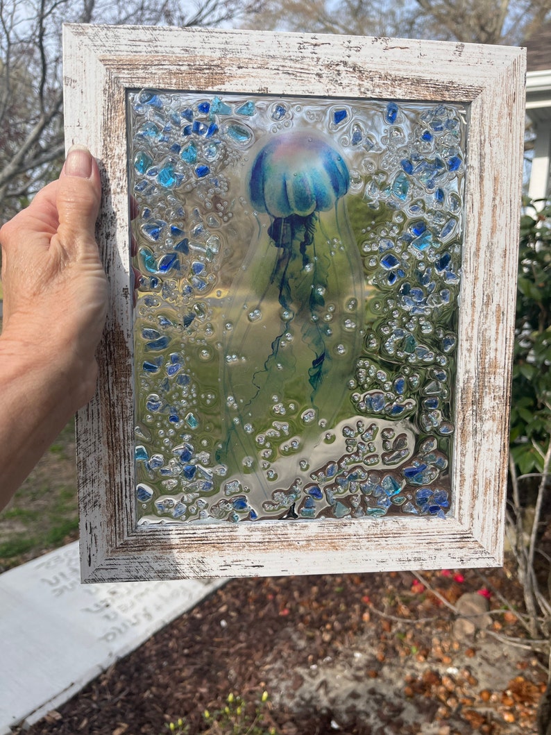 May include: A framed glass art piece featuring a blue jellyfish with a pink bell, surrounded by blue and clear glass pieces. The frame is white with a distressed finish.