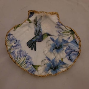 May include: A white seashell with a gold rim, decorated with a blue hummingbird and blue and white flowers.