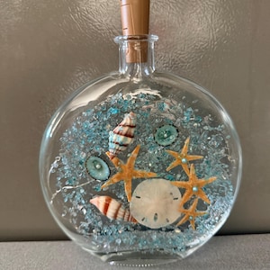May include: A clear glass bottle with a cork stopper, filled with blue decorative stones, seashells, and orange starfish. The bottle has a round shape and is designed to be decorative.
